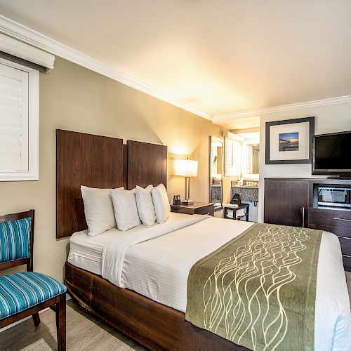 A modern hotel room featuring a bed with multiple pillows, a chair, a TV on a dresser, a window, and vibrant wall art.