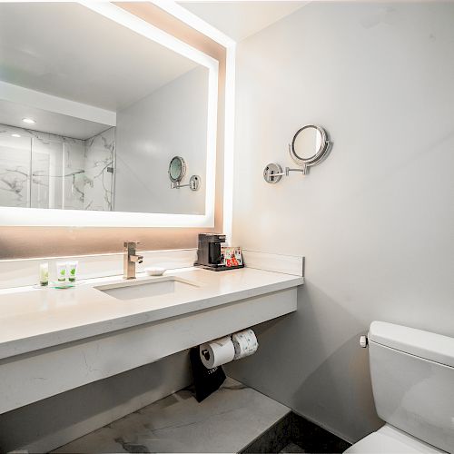 The image shows a modern bathroom with a lit mirror, a washbasin, a toilet, and a shower area in the background. The area is clean and well-organized.