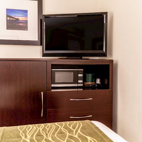 A hotel room has a TV on a wooden dresser, a microwave in the cabinet below, a framed photo on the wall, and part of a bed with a patterned cover.