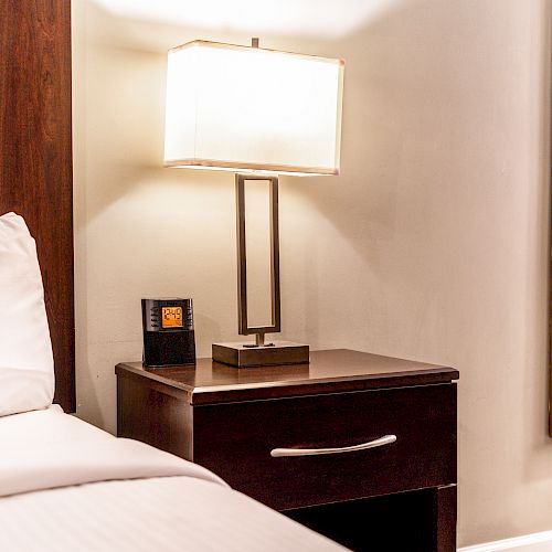 A neatly made bed next to a wooden nightstand with a lamp, digital clock, and a mirror reflecting part of a bathroom.