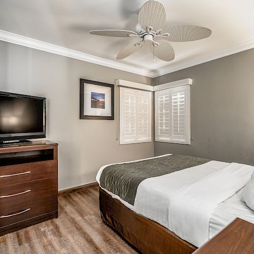 A small, modern bedroom with a bed, a TV on a wooden stand, a ceiling fan, framed art, and light coming through window shutters, ending the sentence.