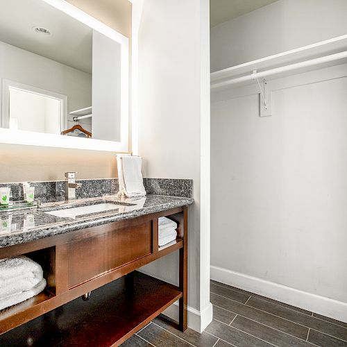 A bathroom with a counter, large mirror, and towels, next to an empty closet with hangers and a blue towel on the right side.