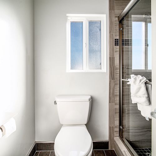 The image shows a clean, modern bathroom with a toilet, a roll of toilet paper, and a glass-enclosed shower next to a small, frosted window.