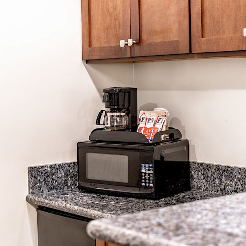 A kitchen counter with a microwave, coffee maker, and several coffee packets, under wooden cabinets, with granite countertops.