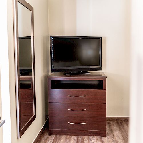 A small room corner featuring a flat-screen TV on a wooden dresser with three drawers, positioned next to a tall mirror on the wall.