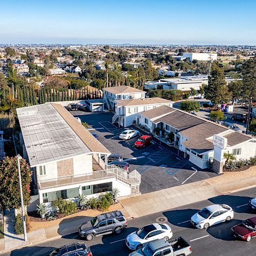 An aerial view of a residential area with multiple buildings, parked cars, and surrounding streets under a clear sky.