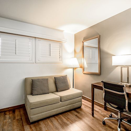 A small room with a tan sofa, desk with chair, floor lamp, table lamp, mirror, and wooden flooring, lit by warm lighting.