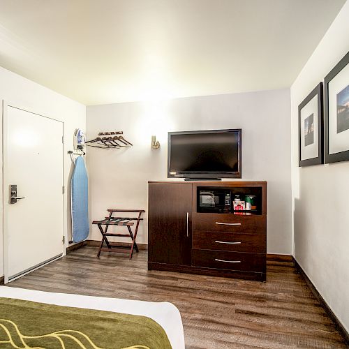 The image shows a hotel room with a TV on a dresser, framed pictures, an ironing board, and a bed in the foreground, featuring a wooden floor.