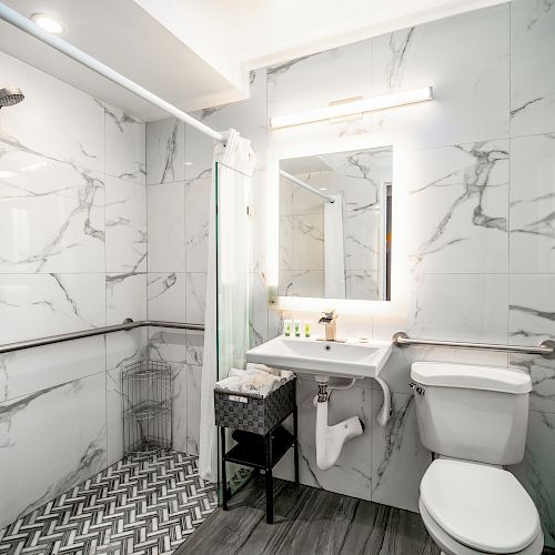 This is a modern bathroom with marble walls, a shower, a toilet, a sink, a lit mirror, and metal grab bars for accessibility.