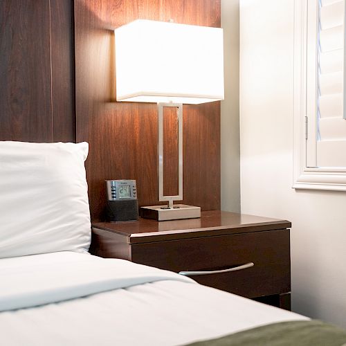 The image shows a neatly made bed beside a wooden nightstand with a lamp, an alarm clock, and window shutters in the background.