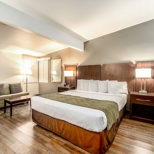 This image shows a modern hotel room with a large bed, nightstands with lamps, a sofa, and a wooden floor. The walls are light gray.