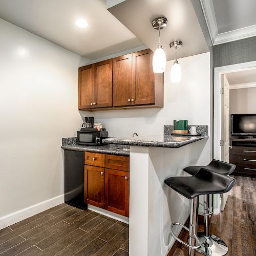 A compact kitchen area with wooden cabinets, bar stools, a sink, microwave, and a mini-fridge next to a small dining table, adjoining another room.