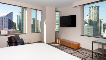 A modern hotel room with city views, featuring a bed, chair, wall-mounted TV, and minimalist decor.