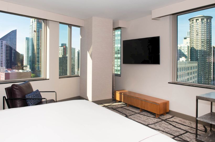 A modern hotel room with city views, featuring a bed, chair, wall-mounted TV, and minimalist decor.