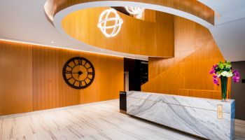 Modern lobby with spiral wooden staircase, marble reception desk, large wall clock, and hanging spherical lights. Flowers on the desk.