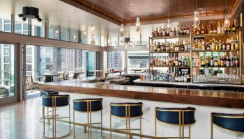 A modern bar with a sleek design features a long wooden counter, high stools, and a wide selection of bottles on shelves. Large windows offer city views.
