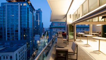 A modern urban balcony with chairs and tables overlooks a city street lined with high-rise buildings during evening twilight.