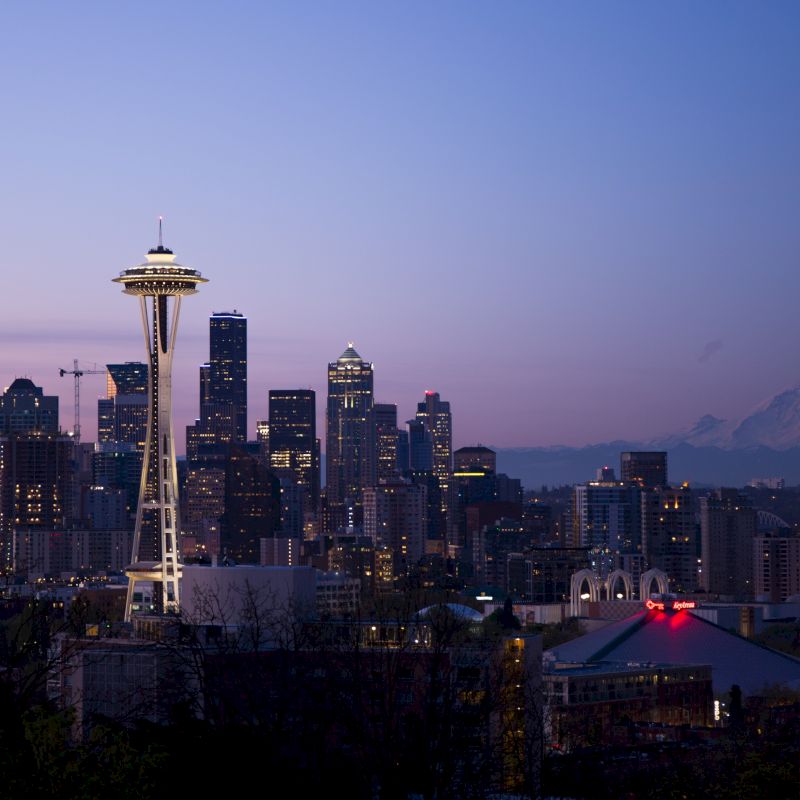 This image shows the Seattle skyline at dusk, featuring the iconic Space Needle with Mount Rainier visible in the background under a twilight sky.
