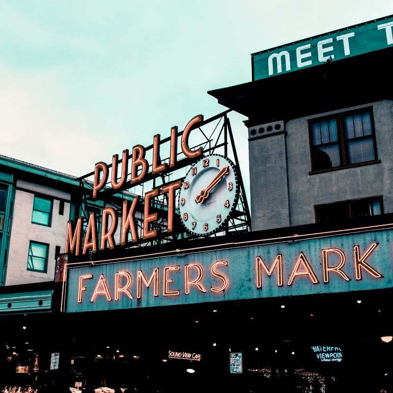 This image shows the entrance of a public market with prominent "Farmers Market" neon signage and a clock. Nearby buildings and signs are visible.