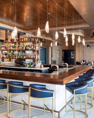 This image shows a modern bar with a wooden countertop, blue barstools, hanging lights, and a well-stocked liquor shelf in a spacious, chic setting.