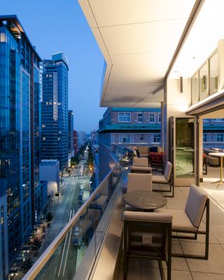 A modern balcony overlooks a city street with tall buildings, featuring comfortable seating and open sliding doors to an interior space.
