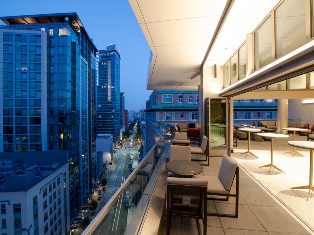 A modern cityscape at dusk with tall buildings and a luxurious balcony featuring chairs and tables, overlooking a well-lit street.