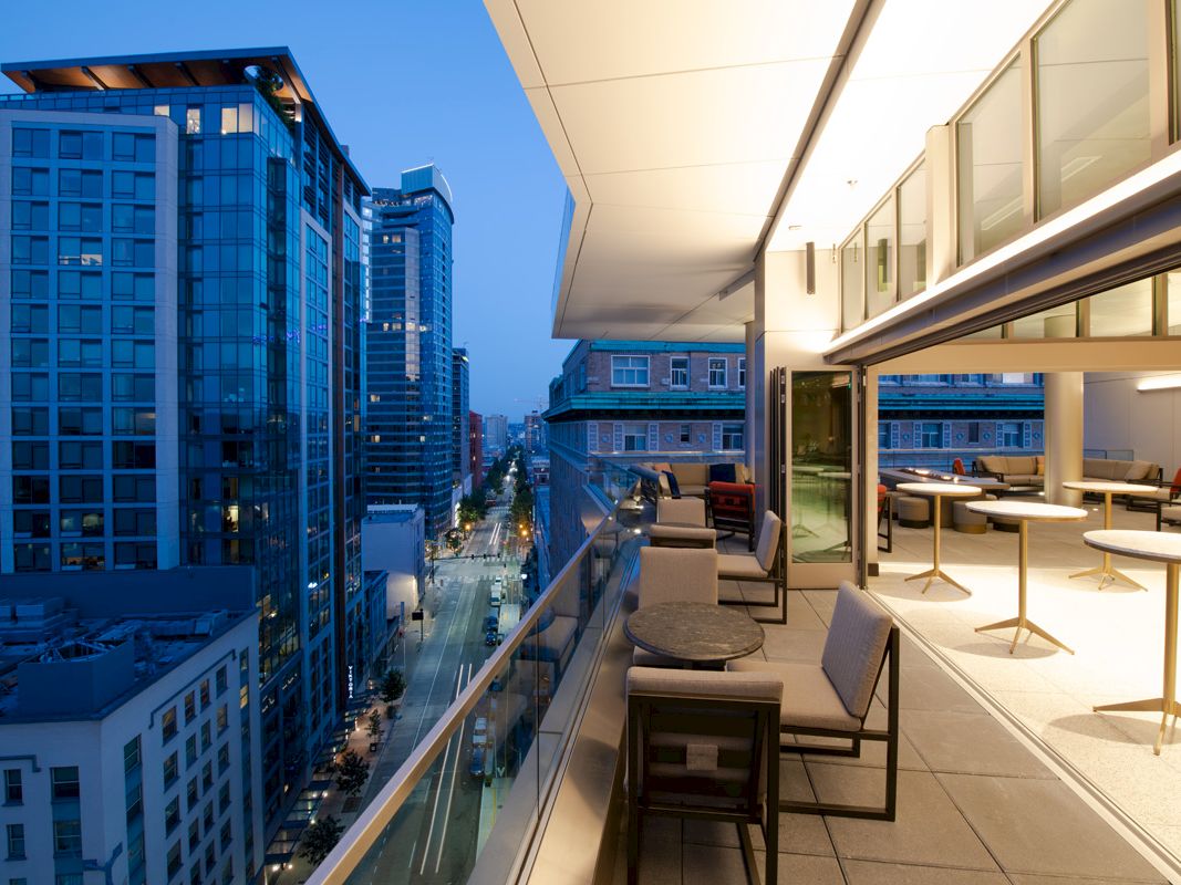 A modern rooftop terrace with seating overlooks a bustling cityscape of tall buildings, lit up as evening approaches. The terrace has tables and chairs.