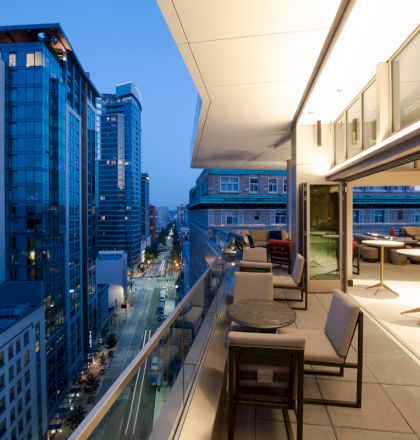 This image shows a modern cityscape with high-rise buildings at dusk, and a rooftop patio with seating and tables offering a view of the street below.