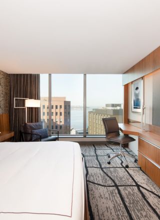 A modern hotel room with a large bed, a desk and chair, a flat-screen TV, and a view of city buildings through a large window.