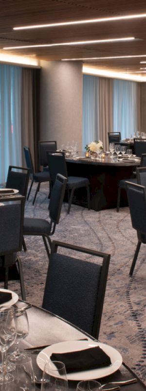 A banquet hall with round tables set with black napkins, plates, glasses, and flowers. The room has dim lighting and sheer curtains.