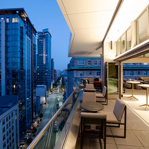 A modern cityscape featuring high-rise buildings is seen from a balcony at dusk, furnished with tables and chairs, and illuminated warmly.