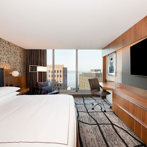 A modern hotel room features a large bed, wall-mounted TV, desk with chair, artistic wallpaper, and a view of buildings through large windows.