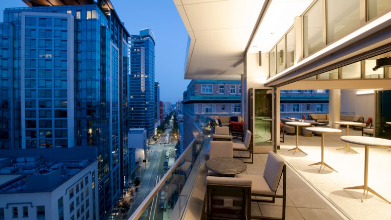 The image shows an elegant rooftop terrace with modern furniture, overlooking a cityscape of tall buildings at dusk, with lights illuminating the streets.