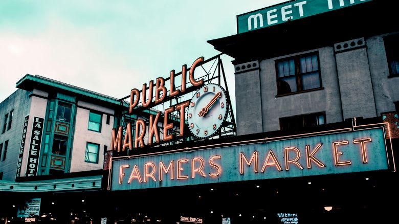 A bustling farmers market is highlighted by a prominent clock and neon signs reading "Public Market" and "Farmers Market," surrounded by older buildings.