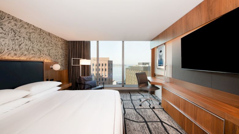 A modern hotel room with a large bed, wall-mounted TV, desk, chair, and floor-to-ceiling windows overlooking city buildings and water.