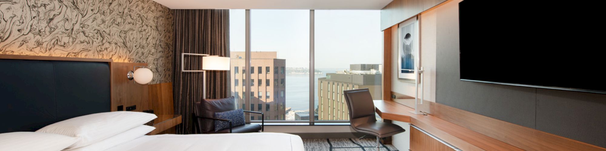 The image shows a modern hotel room with a large bed, a desk with a chair, a flat-screen TV, and a floor-to-ceiling window with a city view.