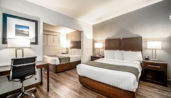 This image shows a modern hotel room with two beds, a work desk with a chair, and two nightstands with lamps, featuring wooden flooring.