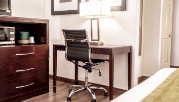 The image shows a modern desk setup with a black office chair, a table lamp, framed wall art, a dresser with drawers, and a glimpse of a bed.