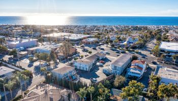 A coastal town with residential buildings, trees, and roads, overlooks the ocean under a clear blue sky with the sun shining on the water.