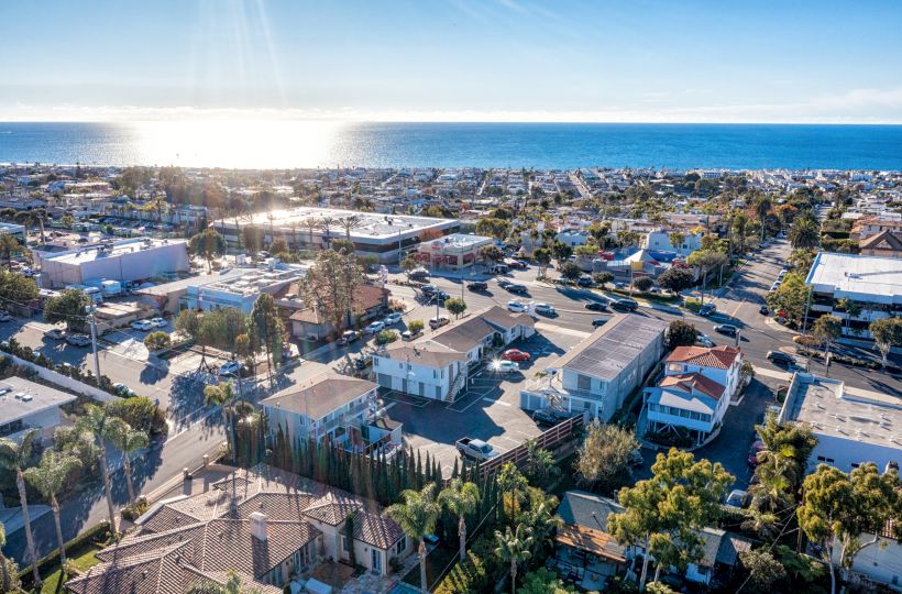 A coastal town with residential buildings, trees, and roads, overlooks the ocean under a clear blue sky with the sun shining on the water.