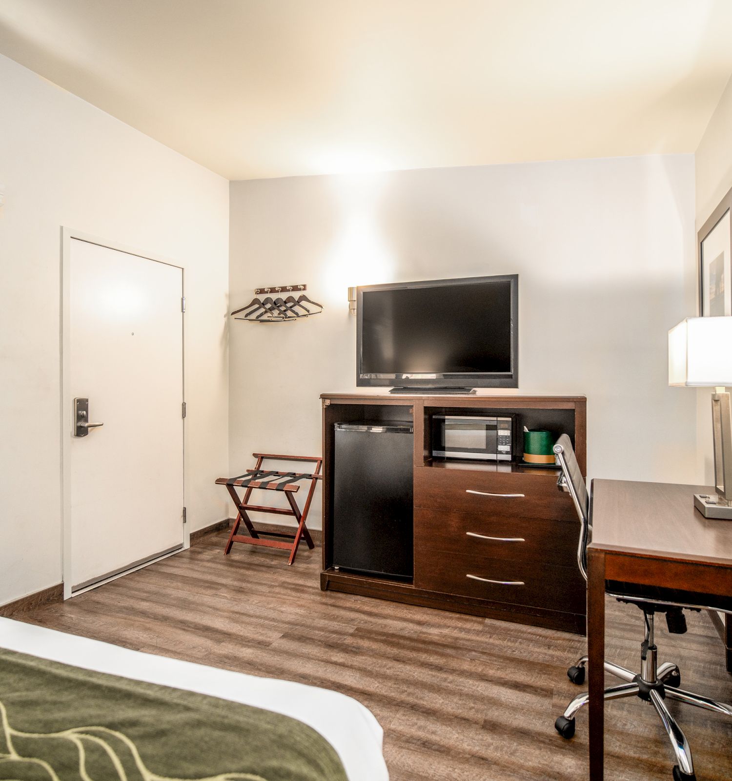 The image shows a hotel room featuring a bed, TV on a cabinet, desk with lamp, microwave, chair, and wall art, with a door and windows.