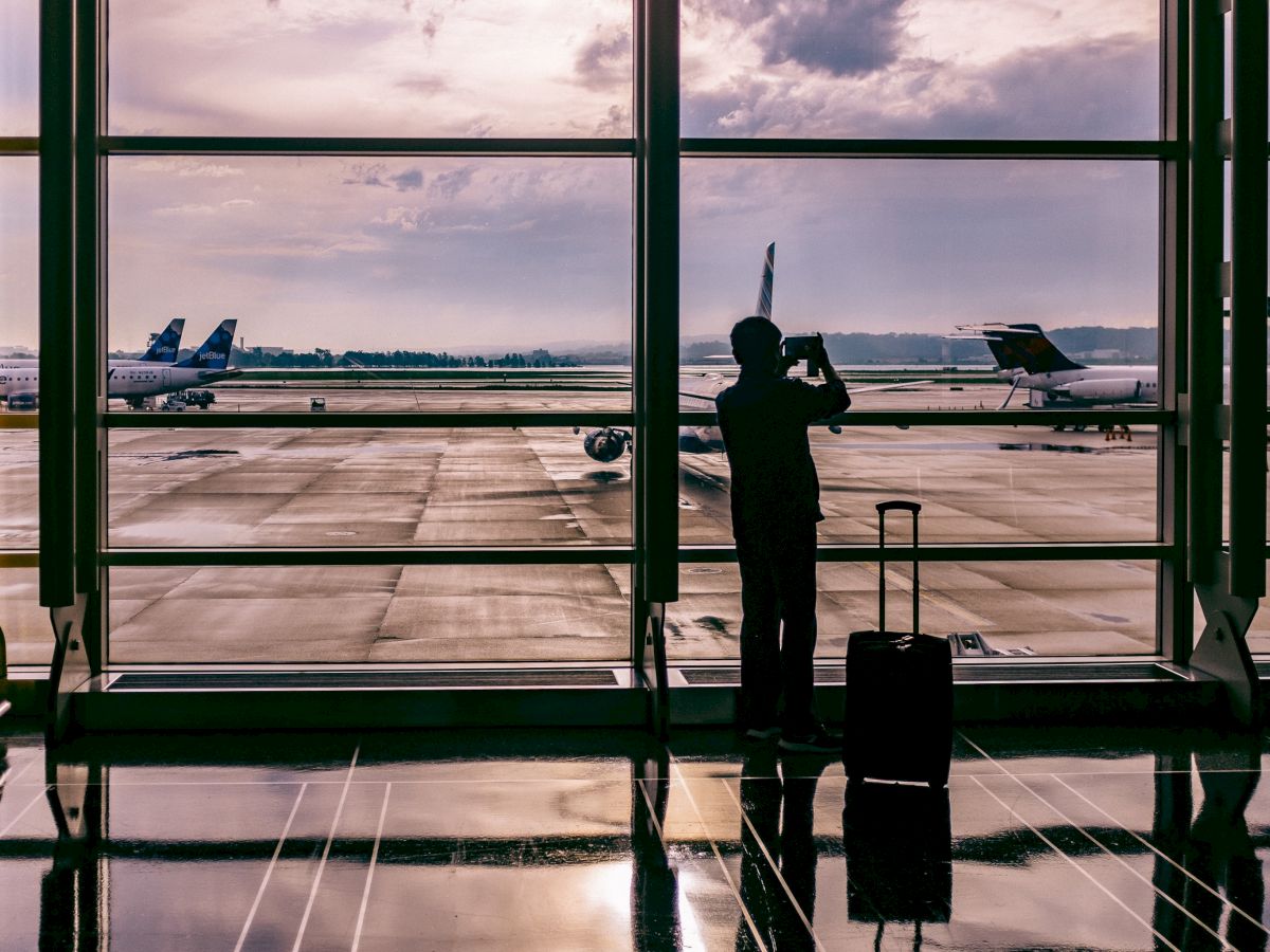 A person with a suitcase is standing in an airport terminal, looking out at several airplanes parked on the runway through large windows.