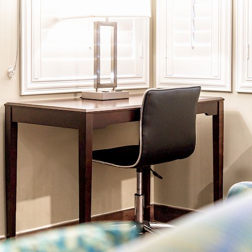 A simple workspace with a dark wooden desk, a modern lamp, a black office chair, and a window with shutters.