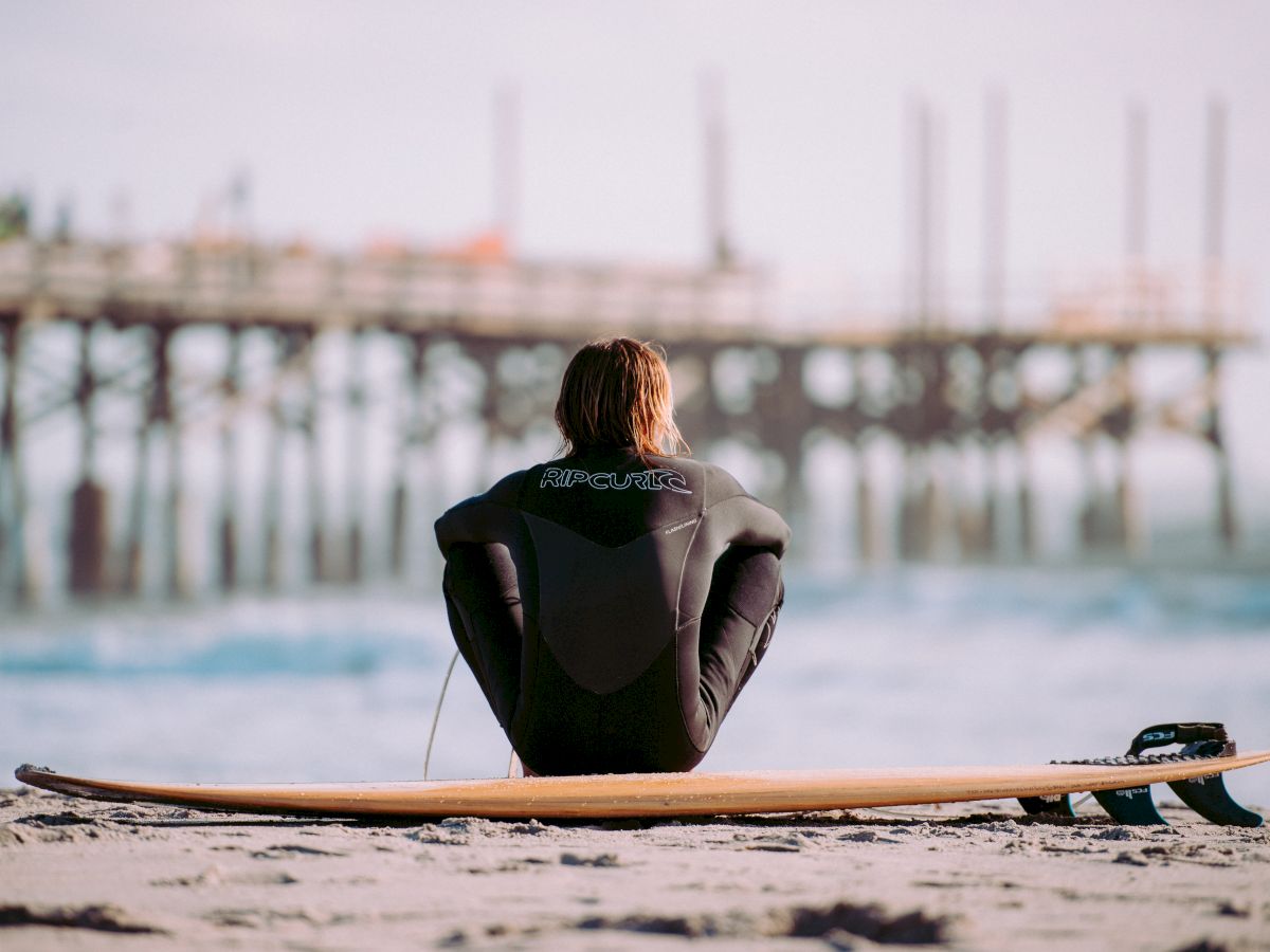 A person in a wetsuit sits on the beach with a surfboard, gazing at the waves with a pier in the background.